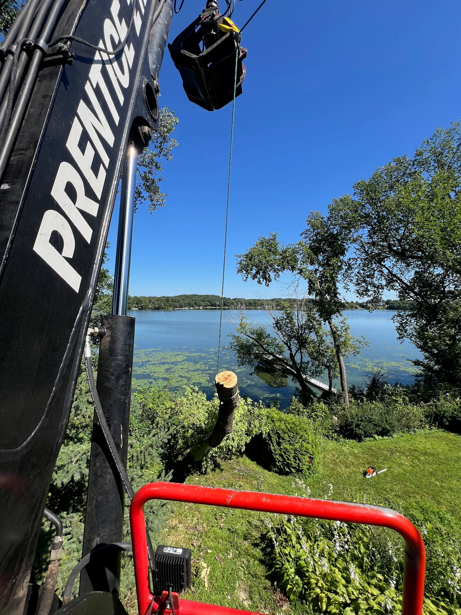 Knuckleboom crane lifting a log section from a wooded hillside above a lake near Dassel, Minnesota